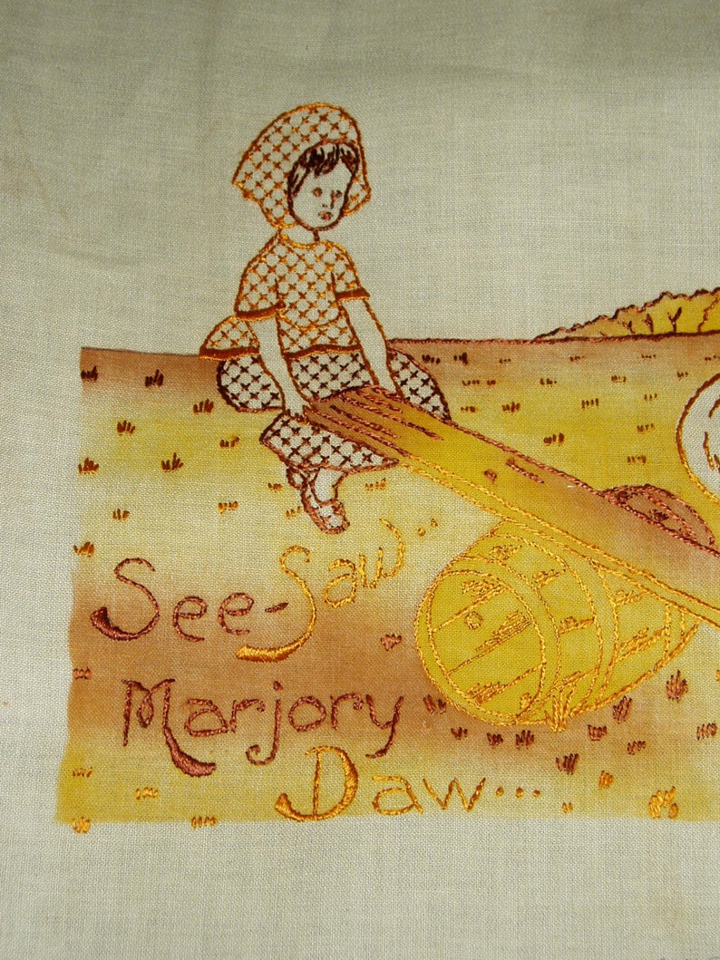 May include: A light-colored fabric panel featuring an embroidered illustration of a child in a checkered outfit sitting on a seesaw. The design includes a wooden barrel support and the embroidered text "See-Saw Marjory Daw..." in brown thread.