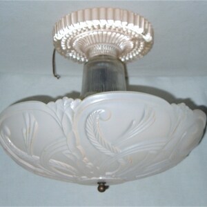 Puede incluir: Una lámpara de techo antigua con una pantalla de vidrio esmerilado. La pantalla tiene un diseño floral ornamentado y un remate decorativo de latón. La lámpara está montada en un techo blanco y probablemente sea de la época Art Déco.