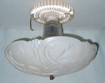 May include: An antique ceiling light fixture with a frosted glass shade. The shade has an ornate floral design and a decorative brass finial. The fixture is mounted on a white ceiling and is likely from the Art Deco era.