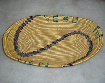 May include: An oval-shaped woven basket with a brown and green design. The basket has a woven handle on each side and the words "YESU" are woven into the design.