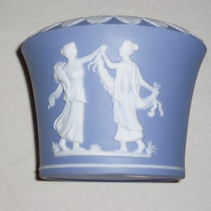 May include: A blue and white ceramic vase with a raised design of two women holding garlands. The vase has a rounded bottom and a flared top.