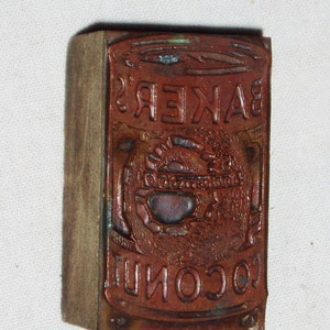 May include: A vintage wooden printing block with a metal plate featuring the text "BAKER'S COCONUT" in a decorative font. The metal plate has a design of a stylized coconut with a sun and leaves.
