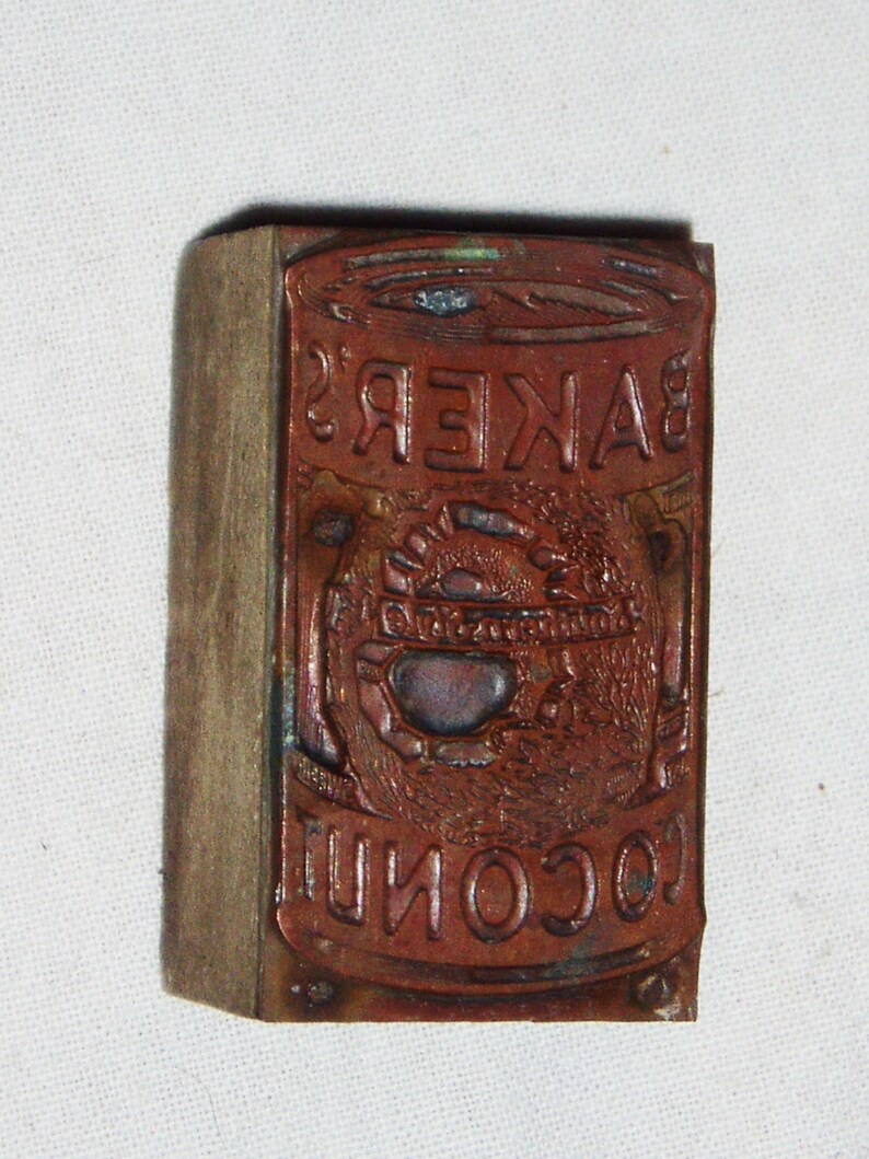May include: A vintage wooden printing block with a metal plate featuring the text "BAKER'S COCONUT" in a decorative font. The metal plate has a design of a stylized coconut with a sun and leaves.