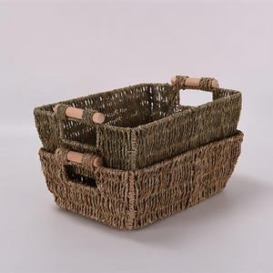 May include: Two rectangular woven baskets stacked on top of each other. The baskets are made of a natural fiber in shades of green and brown. Each basket has a wooden handle on either side.