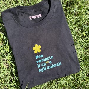 Puede incluir: Camiseta negra con una flor amarilla y el texto italiano "Non rompete il ca**o agli animali" en turquesa. La camiseta tiene cuello redondo y la marca "BRODO + design" en rosa.