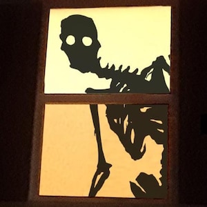 May include: Two rectangular panels with a black skeleton silhouette against a yellow background. The top panel shows the skull and upper body, while the bottom panel displays the lower body and legs. The panels are set within a dark brown frame.