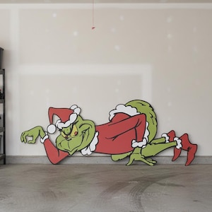 Puede incluir: Un gran recorte de madera colorido del Grinch con un gorro de Papá Noel y un traje rojo, tumbado en el suelo. El Grinch es verde con un ribete blanco en su sombrero y traje.
