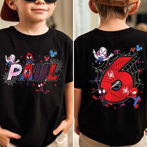 May include: Black t-shirt featuring a colorful Spider-Man design. The front displays the name "PAUL" in a webbed font with Spider-Man characters. The back shows a large red number "6" with a web design and more Spider-Man characters.