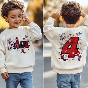 May include: Cream-colored sweatshirt featuring the name "PAUL" in a colorful, web-themed design with Spider-Man graphics. The back of the sweatshirt displays a large red number "4" with Spider-Man and web details, suitable for a child's birthday.