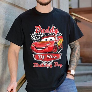 Puede incluir: Camiseta negra con un dise&ntilde;o de Rayo McQueen de la pel&iacute;cula Cars. El dise&ntilde;o incluye el texto "Daddy Of The Birthday Boy" encima y debajo de la imagen del coche, con banderas a cuadros en el fondo.