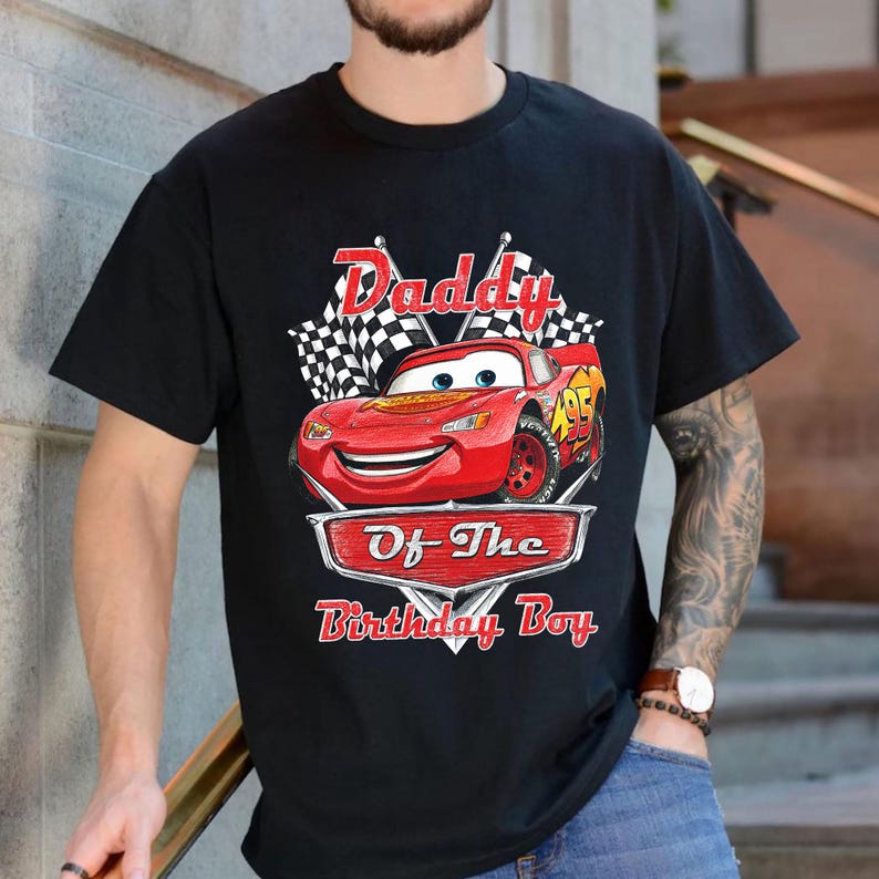 Puede incluir: Camiseta negra con un dise&ntilde;o de Rayo McQueen de la pel&iacute;cula Cars. El dise&ntilde;o incluye el texto "Daddy Of The Birthday Boy" encima y debajo de la imagen del coche, con banderas a cuadros en el fondo.