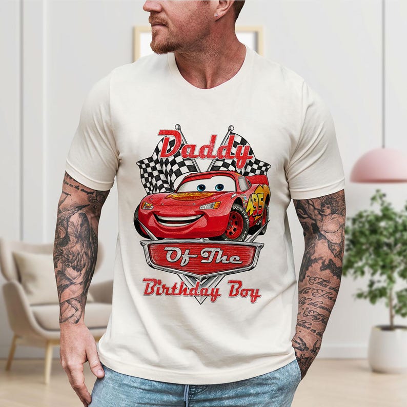 Puede incluir: Camiseta color crema con un gr&aacute;fico de un coche de carreras rojo y el texto "Daddy Of The Birthday Boy". El dise&ntilde;o incluye banderas a cuadros y una fuente estilizada. La camiseta es de manga corta.