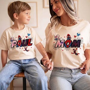 May include: Two matching beige t-shirts with the words "PAUL" and "MOM" in a colorful, comic book style. Each shirt features Spider-Man graphics and web designs. The shirts are worn by a child and an adult.