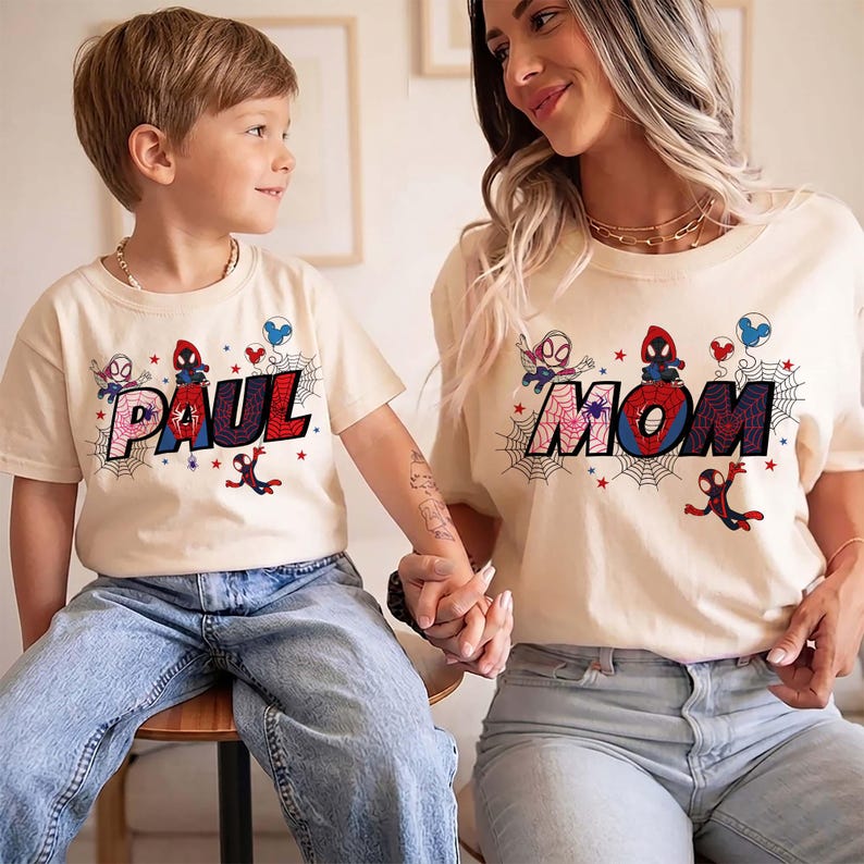 May include: Two matching beige t-shirts with the words "PAUL" and "MOM" in a colorful, comic book style. Each shirt features Spider-Man graphics and web designs. The shirts are worn by a child and an adult.