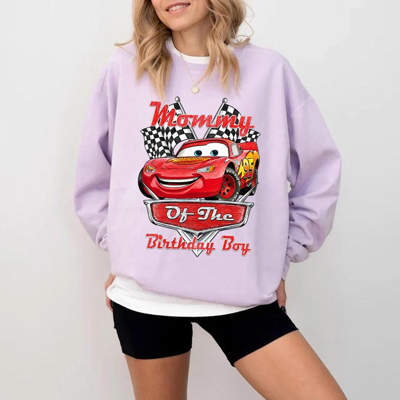Puede incluir: Una sudadera lila con un gr&aacute;fico de un coche de carreras rojo y el texto "Mommy Of The Birthday Boy". El dise&ntilde;o incluye banderas a cuadros y un emblema en forma de escudo. La sudadera es de estilo casual.