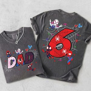 May include: Two gray t-shirts with Spiderman graphics. One shirt says "DAD" with a spiderweb design. The other shirt has a large red number "6" with a spiderweb and Spiderman characters. Both shirts have a chain necklace.