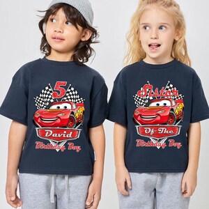 Puede incluir: Dos camisetas azul marino con un dise&ntilde;o de coche de carreras rojo de dibujos animados. Una camiseta dice "5" y "David Birthday Boy", la otra dice "Sister Of The Birthday Boy". Ambas tienen un dise&ntilde;o de bandera a cuadros.