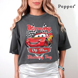 Puede incluir: Camiseta gris oscuro con un coche de carreras rojo de dibujos animados y el texto "Mommy Of The Birthday Boy". El dise&ntilde;o incluye banderas a cuadros y una pancarta roja. La camiseta es de corte holgado.