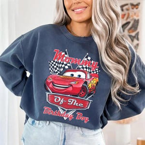 Puede incluir: Sudadera azul oscuro con un coche de carreras rojo de dibujos animados con el n&uacute;mero 95. El texto "Mommy Of The Birthday Boy" est&aacute; impreso encima y debajo del coche en rojo. Banderas a cuadros a ambos lados del coche.