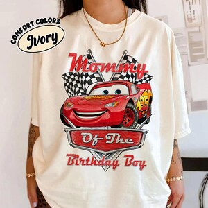Puede incluir: Camiseta color marfil con un coche de carreras rojo de dibujos animados y el texto "Mommy Of The Birthday Boy". El dise&ntilde;o incluye banderas a cuadros y un emblema en forma de escudo. La camiseta es de la colecci&oacute;n Comfort Colors.