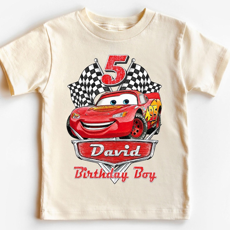 Puede incluir: Camiseta color crema con un dise&ntilde;o del personaje Rayo McQueen de la pel&iacute;cula Cars. El dise&ntilde;o incluye el n&uacute;mero 5, banderas a cuadros y el texto "David Birthday Boy" en rojo.