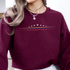 May include: A maroon long-sleeve crewneck sweatshirt featuring a graphic design on the chest. The design consists of a horizontal row of eight white stars above three thin parallel lines in yellow, blue, and red colors.