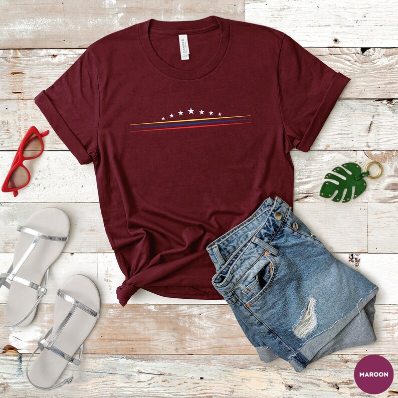 May include: A maroon short-sleeve t-shirt featuring a graphic of seven white stars arranged in an arc above three thin horizontal lines in yellow, blue, and red. The shirt is displayed alongside red sunglasses, blue denim shorts, and a green leaf keychain.