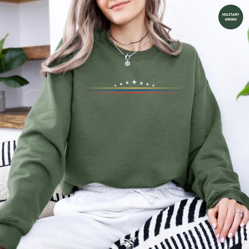 May include: A military green long-sleeve sweatshirt featuring a graphic design on the chest. The design consists of a horizontal arc of eight white stars positioned above three thin, parallel horizontal lines in yellow, blue, and red colors.