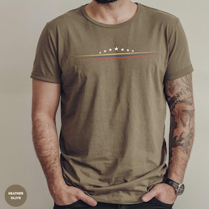 May include: An olive green short-sleeve t-shirt featuring a graphic design on the chest. The design consists of seven white stars arranged in an arc above three thin horizontal lines in yellow, blue, and red colors across the center.