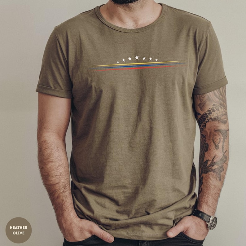 May include: An olive green short-sleeve t-shirt featuring a graphic design on the chest. The design consists of seven white stars arranged in an arc above three thin horizontal lines in yellow, blue, and red colors across the center.