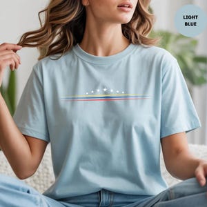May include: A light blue t-shirt featuring a graphic design of eight white stars arranged in an arc above three horizontal lines in yellow, blue, and red. The shirt has a crew neckline and short sleeves, with the text LIGHT BLUE in a circle.