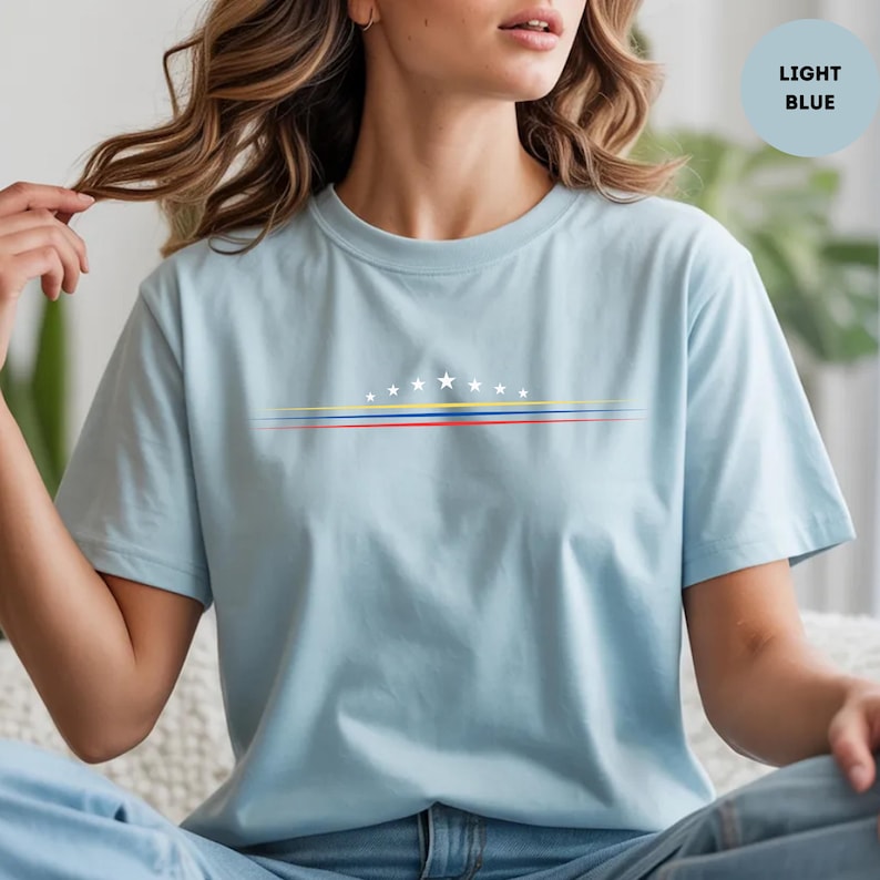 May include: A light blue t-shirt featuring a graphic design of eight white stars arranged in an arc above three horizontal lines in yellow, blue, and red. The shirt has a crew neckline and short sleeves, with the text LIGHT BLUE in a circle.