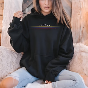 May include: A black hooded sweatshirt featuring a graphic design on the chest with seven white stars arranged in an arc above three thin horizontal lines in yellow, blue, and red. The hoodie includes a front kangaroo pocket and long sleeves.