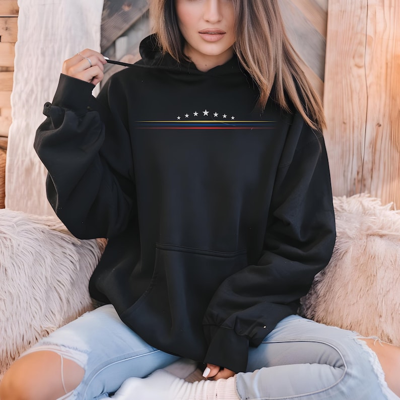 May include: A black hooded sweatshirt featuring a graphic design on the chest with seven white stars arranged in an arc above three thin horizontal lines in yellow, blue, and red. The hoodie includes a front kangaroo pocket and long sleeves.