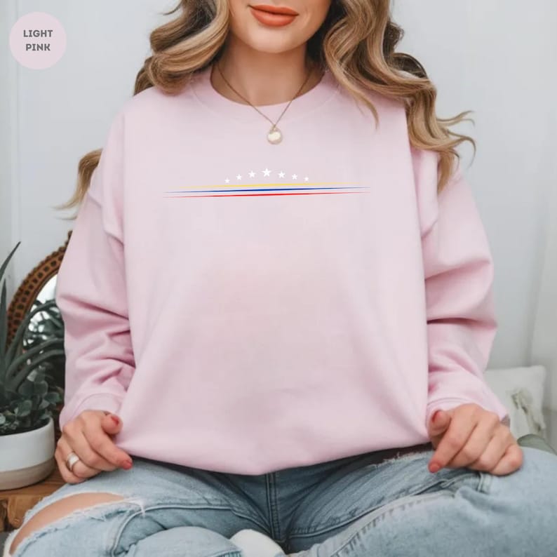 May include: A light pink crewneck sweatshirt featuring a graphic on the chest with eight white stars arranged in an arc above three horizontal lines in yellow, blue, and red. The sweatshirt has long sleeves and a ribbed collar, cuffs, and hem.