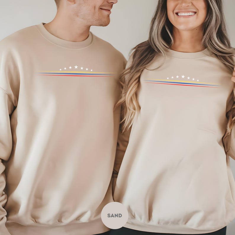 May include: A sand-colored crewneck sweatshirt featuring a graphic on the chest. The design consists of seven white stars arranged in an arc above three horizontal lines in yellow, blue, and red. The sweatshirt has long sleeves and a ribbed collar.