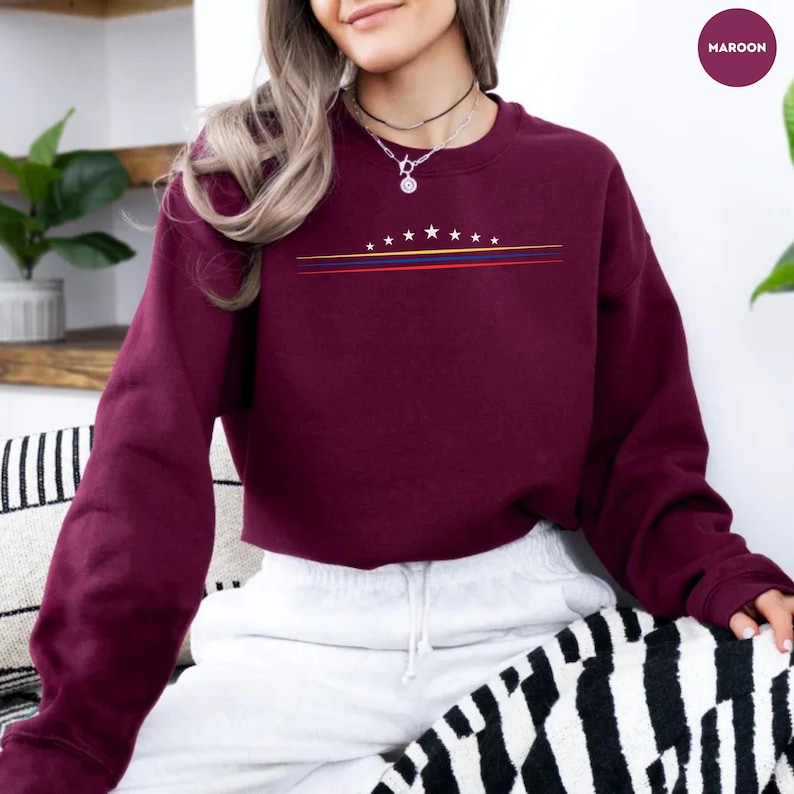 May include: A maroon long-sleeve crewneck sweatshirt featuring a graphic design on the chest. The design consists of a horizontal row of eight white stars above three thin parallel lines in yellow, blue, and red colors.