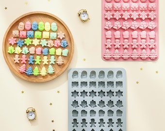 May include: A round wooden tray containing various colorful holiday-themed shapes, a pink silicone mold, and a light blue silicone mold. The molds feature cavities shaped like Santa faces, gift boxes, snowflakes, bells, mittens, gingerbread men, and trees.