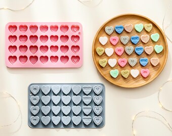 May include: A rectangular pink silicone mold with 35 heart-shaped cavities, a rectangular gray silicone mold with 35 heart-shaped cavities, and a round wooden tray containing 35 small, multi-colored heart-shaped candies with various textures.