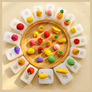 May include: A circular wooden tray surrounded by white silicone moulds, each containing a miniature fruit-shaped eraser. The collection includes colourful representations of grapes, bananas, lemons, strawberries, oranges, and peaches.
