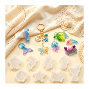 May include: A collection of sea-themed resin charms including a seahorse, mermaid tail, turtle, jellyfish, crab, and dolphin, alongside a gold-toned keychain with anchor and starfish charms, plus eight clear silicone moulds for crafting.