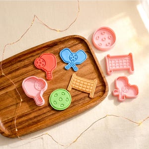 May include: A collection of plastic cookie cutters in various shapes including pickleball paddles, a pickleball, and a net. The cutters are coloured red, blue, light pink, green, and orange. They are arranged on a wooden tray with string lights.