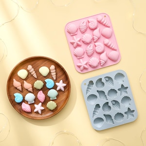 May include: A wooden plate with seashell-shaped sweets in pastel shades, next to two silicone moulds filled with seashell shapes. The moulds are pink and blue, ideal for making themed treats.