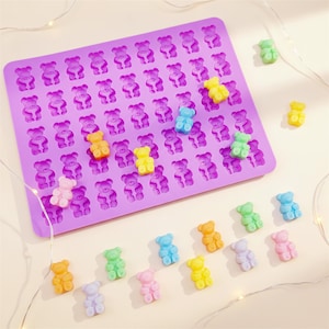 May include: A purple silicone gummy bear mould filled with colourful gummy bears. The mould has multiple bear-shaped cavities. Scattered around the mould are individual gummy bears in various colours, including yellow, green, and pink.