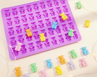 May include: A purple silicone gummy bear mold filled with colorful gummy bears. The mold has multiple bear-shaped cavities. Scattered around the mold are individual gummy bears in various colors, including yellow, green, and pink.