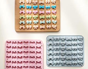 May include: An overhead shot displays three bow-shaped molds. One is a wooden cutting board with colorful bows. The other two are silicone molds, one pink and one blue, each filled with bow-shaped cavities. These are likely for crafting or baking.