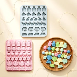 Peut inclure: Trois moules en silicone et un plateau en bois remplis de bonbons sur le thème du basket-ball. Les moules sont bleu clair et roses, chacun avec diverses formes comme des maillots, des trophées, des ballons et des paniers. Le plateau contient des bonbons colorés de formes similaires.