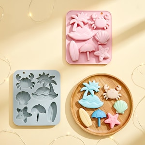 Peut inclure: Trois moules en silicone et un plateau en bois avec des formes sur le thème de la plage. Les moules sont roses et bleus, avec des palmiers, des crabes, des coquillages, des planches de surf, des parapluies et des étoiles de mer. Le plateau en bois contient les mêmes formes.