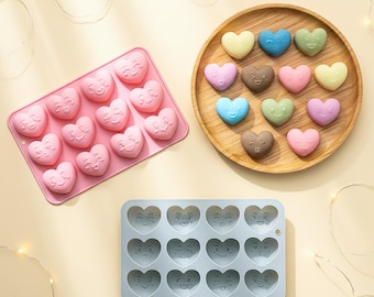 May include: A pink silicone mold with twelve heart-shaped cavities, a light blue silicone mold with twelve heart-shaped cavities, and a round wooden plate holding fourteen small, colorful heart-shaped items with various facial expressions.