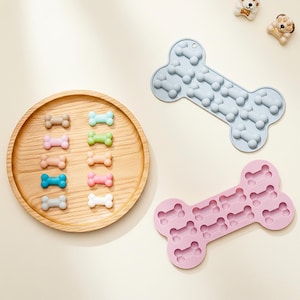 May include: A wooden tray displays colourful dog bone-shaped treats. Two silicone moulds, one light blue and one pink, are shaped like large dog bones with bone-shaped cavities. Two small dog-shaped decorations are in the upper right corner. The items are on a light beige surface.